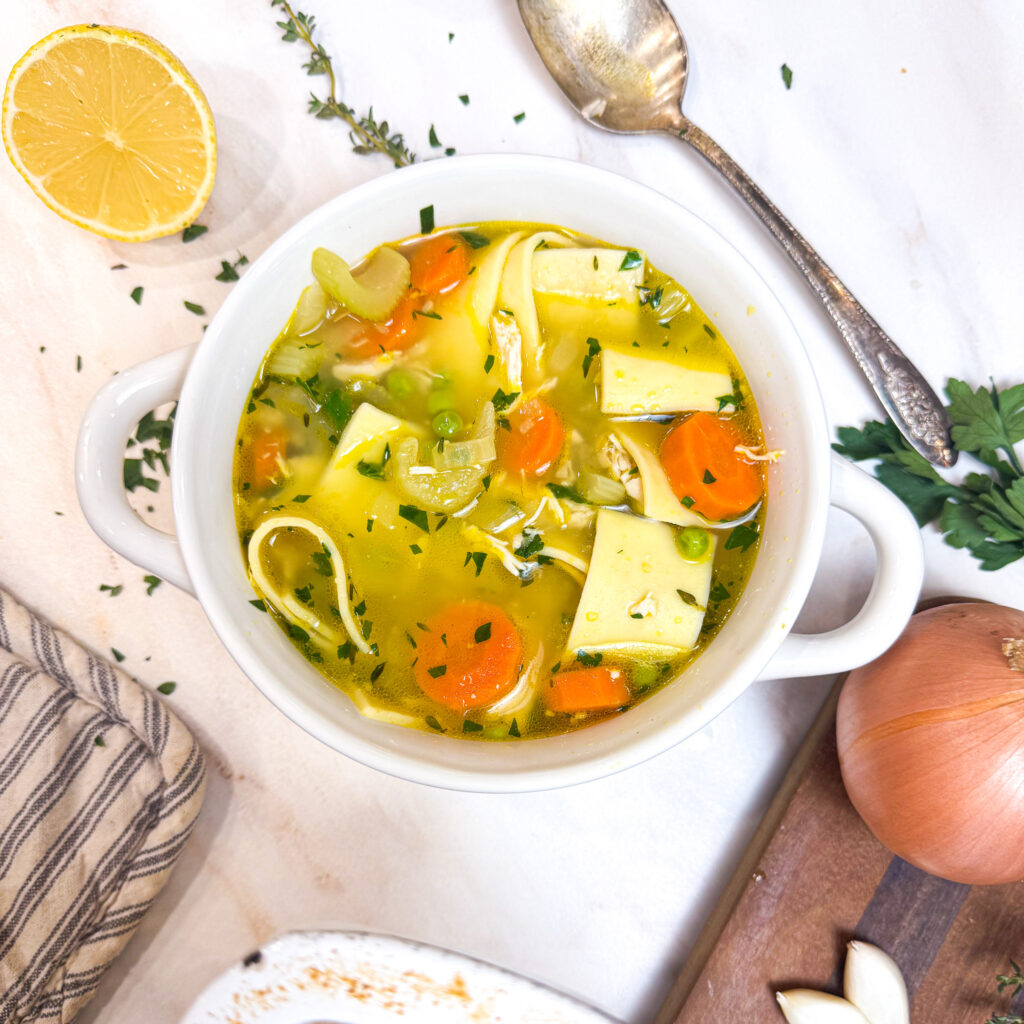 A cozy bowl of homemade chicken noodle soup filled with wide egg noodles, tender chicken, carrots, celery, peas, and fresh herbs in a golden broth. A halved lemon, fresh thyme, and parsley sit nearby on a marble surface, adding a bright, comforting touch to the scene.