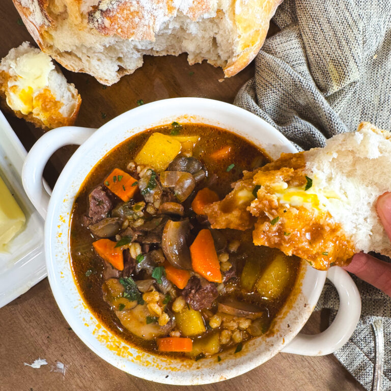A hand dipping a buttery piece of rustic bread into a bowl of beef barley mushroom soup. The bowl is filled with tender veggies, barley, and beef, and a loaf of crusty bread sits in the background.