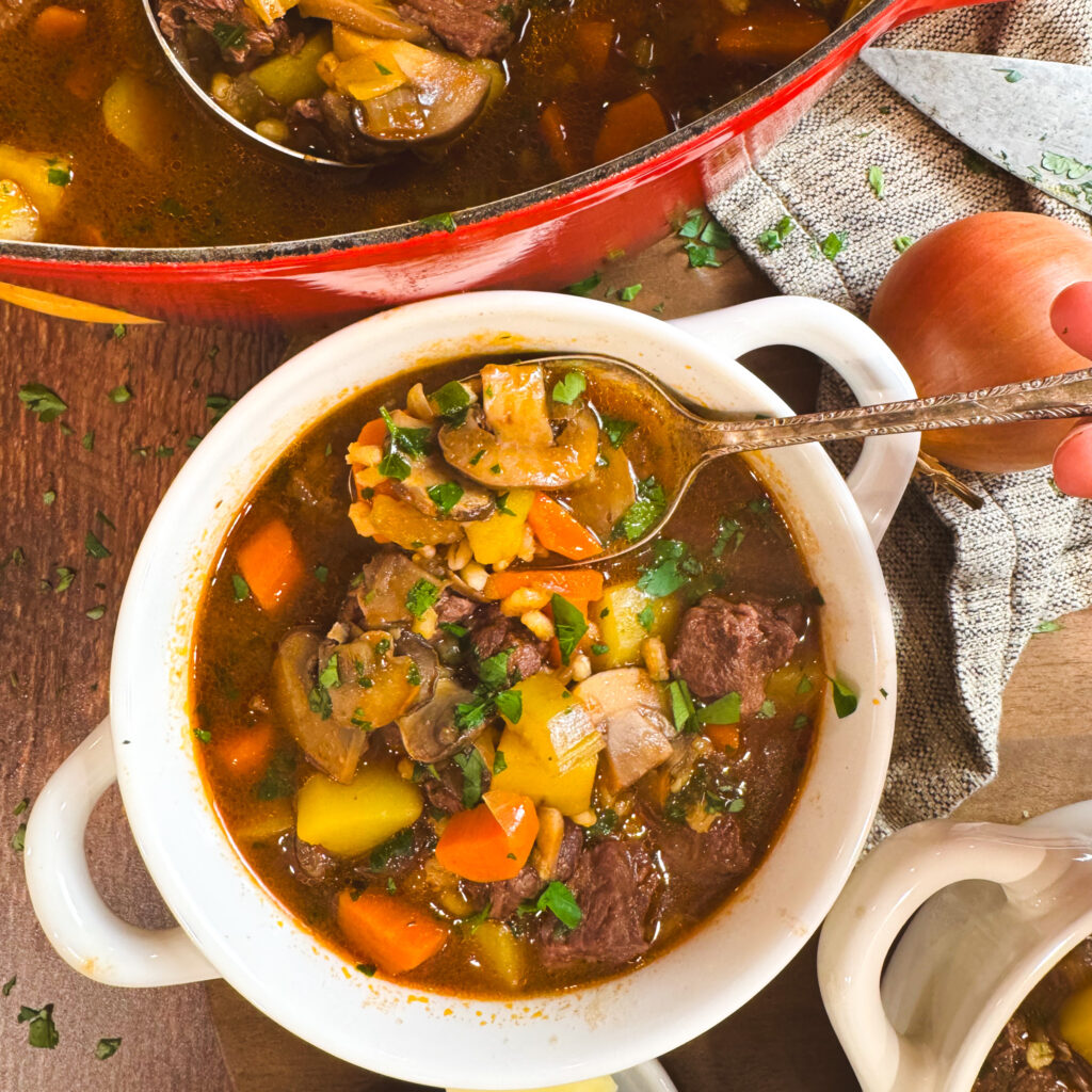 A white soup bowl filled with beef barley mushroom soup, with a ladle lifting a scoop from the red Dutch oven behind it. The bowl is topped with colorful vegetables, mushrooms, barley, and beef, garnished with parsley.