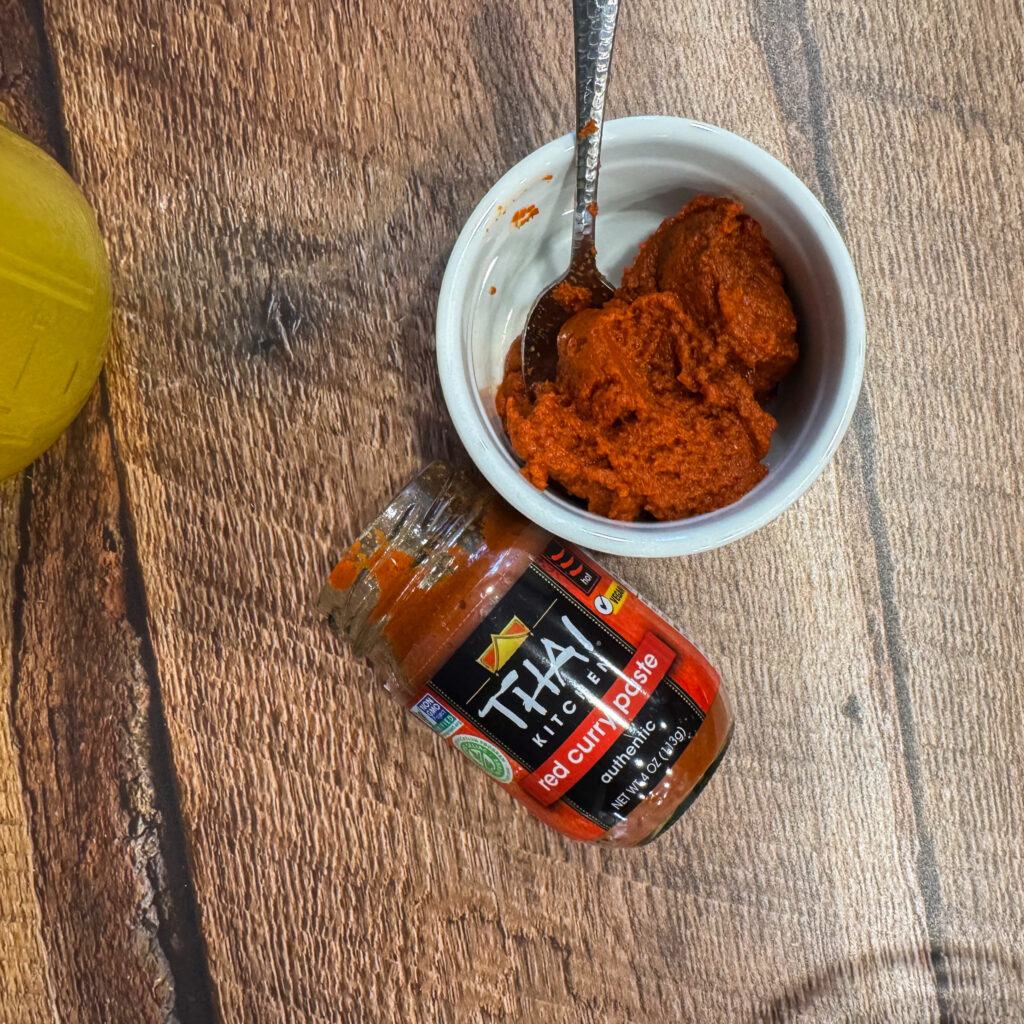 Spoonful of vibrant red curry paste beside an open jar of Thai Kitchen curry paste on a wooden countertop.