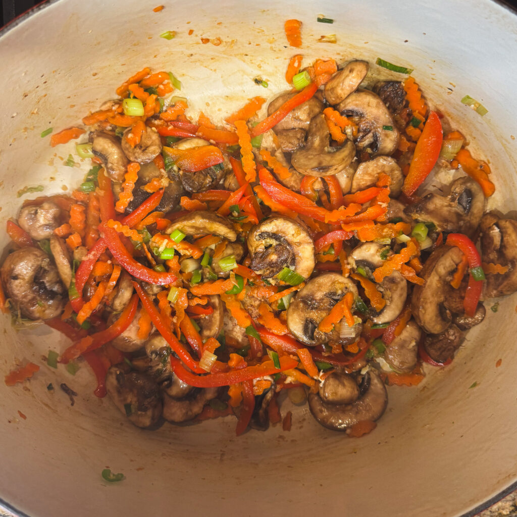 Sautéed mushrooms, carrots, and red bell peppers in a Dutch oven, creating a colorful base for creamy curry noodles.