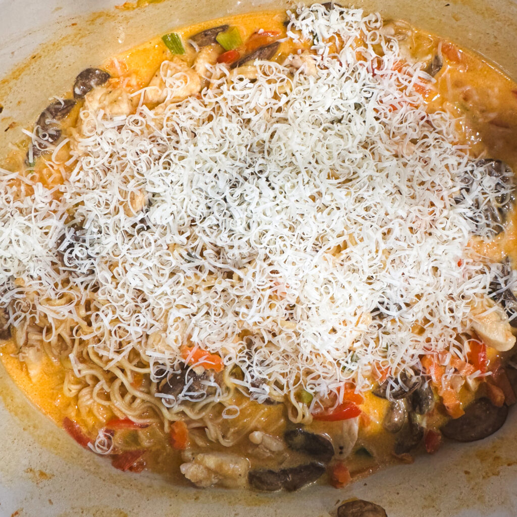 Creamy curry ramen in a Dutch oven topped generously with freshly grated Parmesan, ready to melt into the sauce.