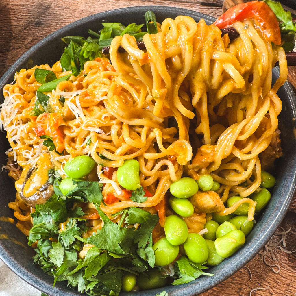 Chopsticks lifting a swirl of saucy curry noodles from a bowl filled with chicken, mushrooms, and edamame — creamy curry coating every bite.