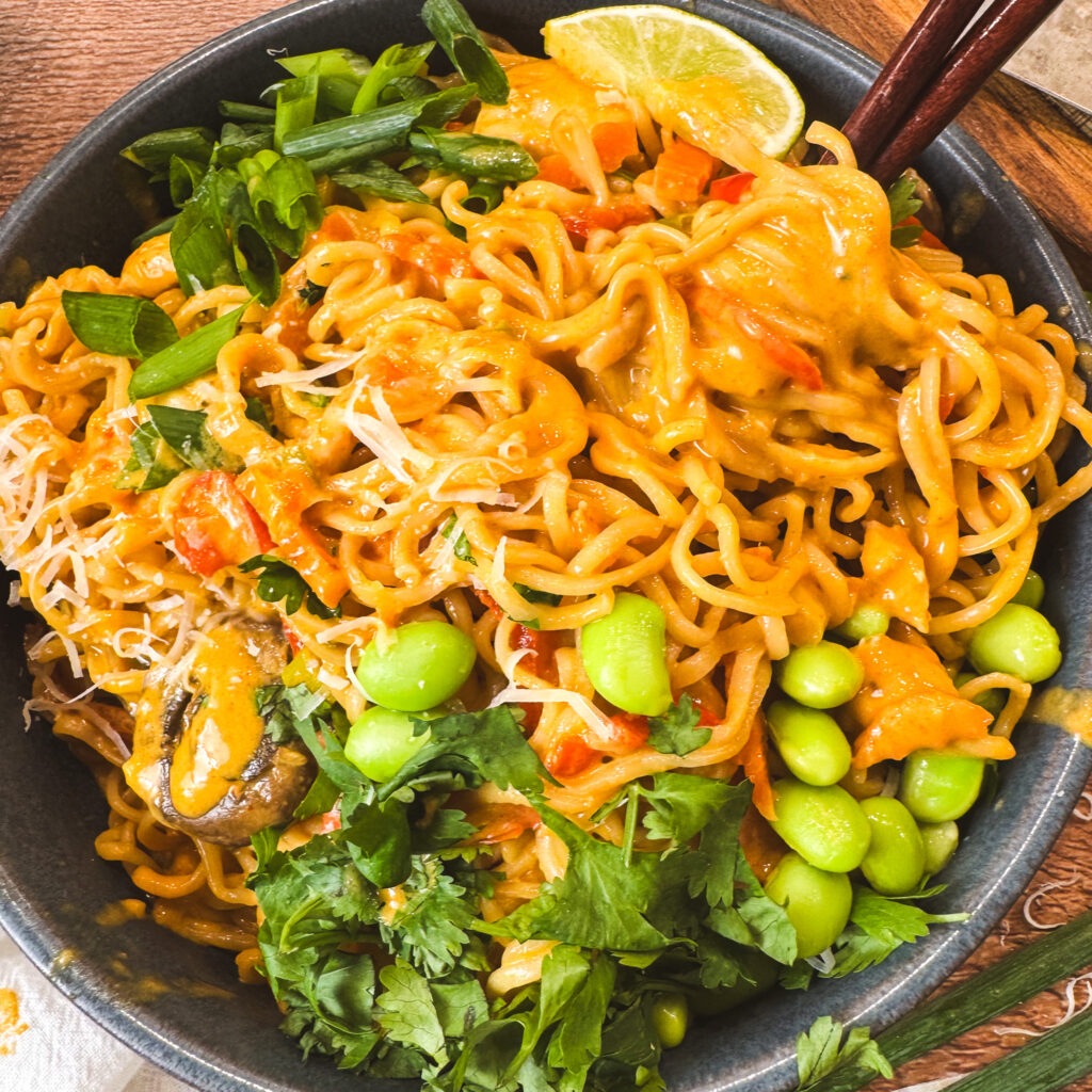 Close-up of a bowl of creamy curry ramen noodles topped with bright green edamame, fresh cilantro, sliced green onions, and a lime wedge — golden curry sauce clings to every noodle.