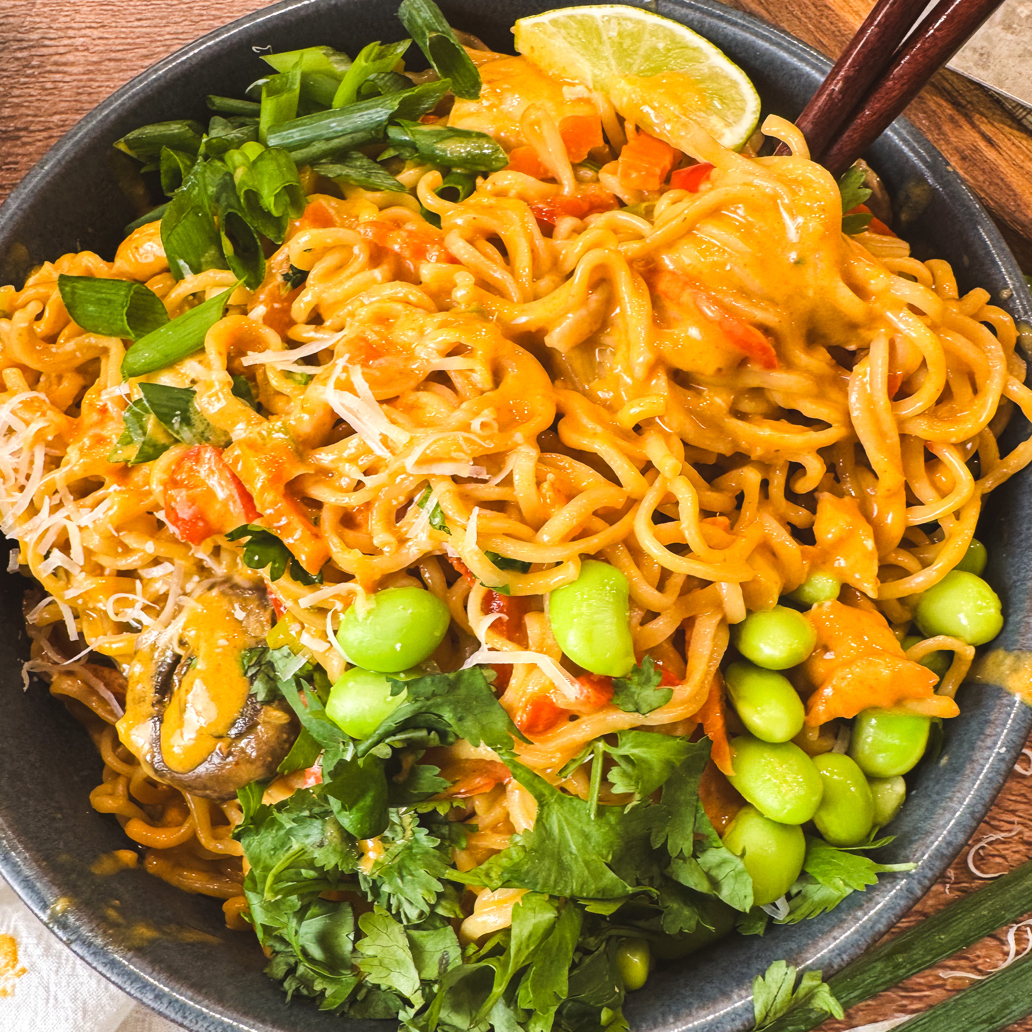 Close-up of a bowl of creamy curry ramen noodles topped with bright green edamame, fresh cilantro, sliced green onions, and a lime wedge — golden curry sauce clings to every noodle.