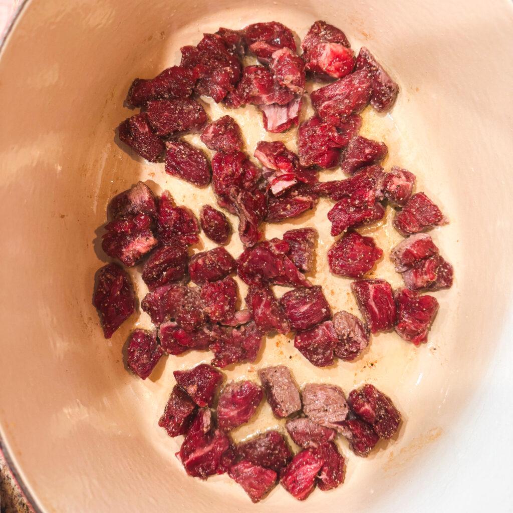 A Dutch oven with a single layer of raw, seasoned beef cubes beginning to sear. The pieces are spaced out so they brown properly, sizzling lightly in a thin layer of oil.