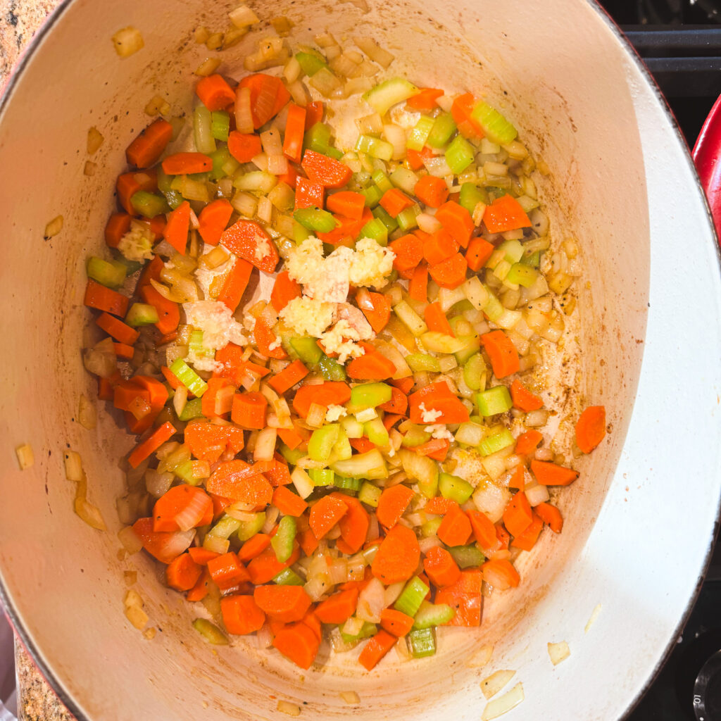 A Dutch oven filled with sautéed onions, celery, and carrots that have started to soften. A small mound of freshly minced garlic sits in the center, ready to be stirred in.