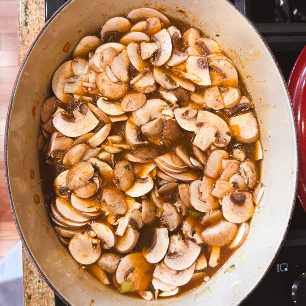 A generous layer of sliced cremini mushrooms added into the pot on top of the tomato-coated vegetables, beginning to soften and release their juices into the broth.