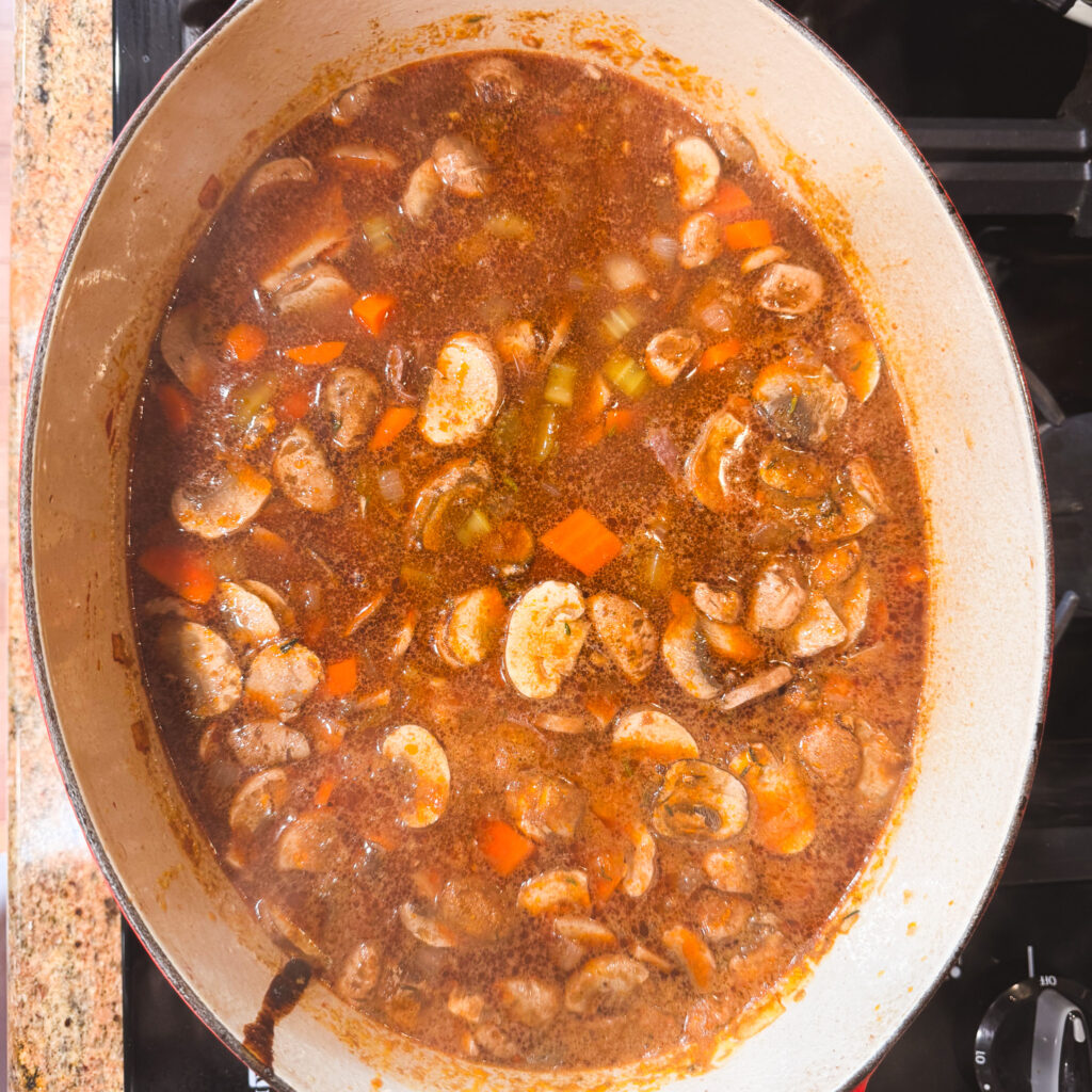 The pot now filled with broth, mushrooms, and vegetables, gently simmering. Bubbles rise to the surface and the broth has turned a warm brown color from the tomato paste and mushroom juices.