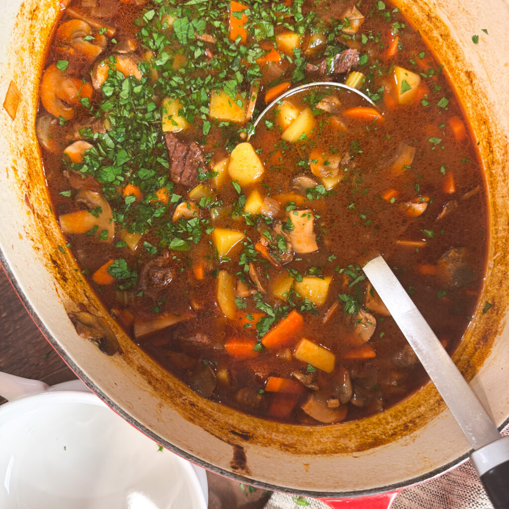 A large Dutch oven filled with hearty beef barley mushroom soup, packed with tender chunks of beef, carrots, celery, potatoes, and mushrooms. Fresh parsley is sprinkled over the top, and a ladle rests inside, ready to serve.