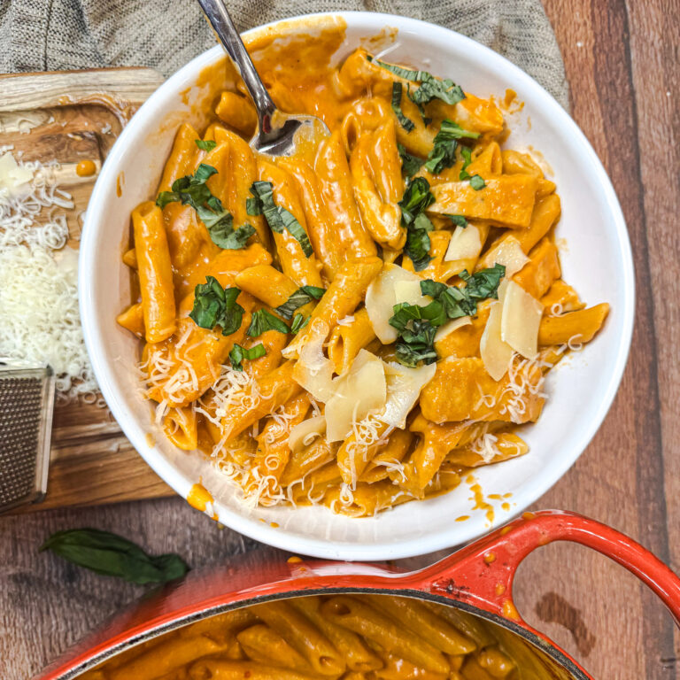 Creamy penne alla vodka in a white bowl topped with ribbons of parmesan and chopped basil for a bright finish.