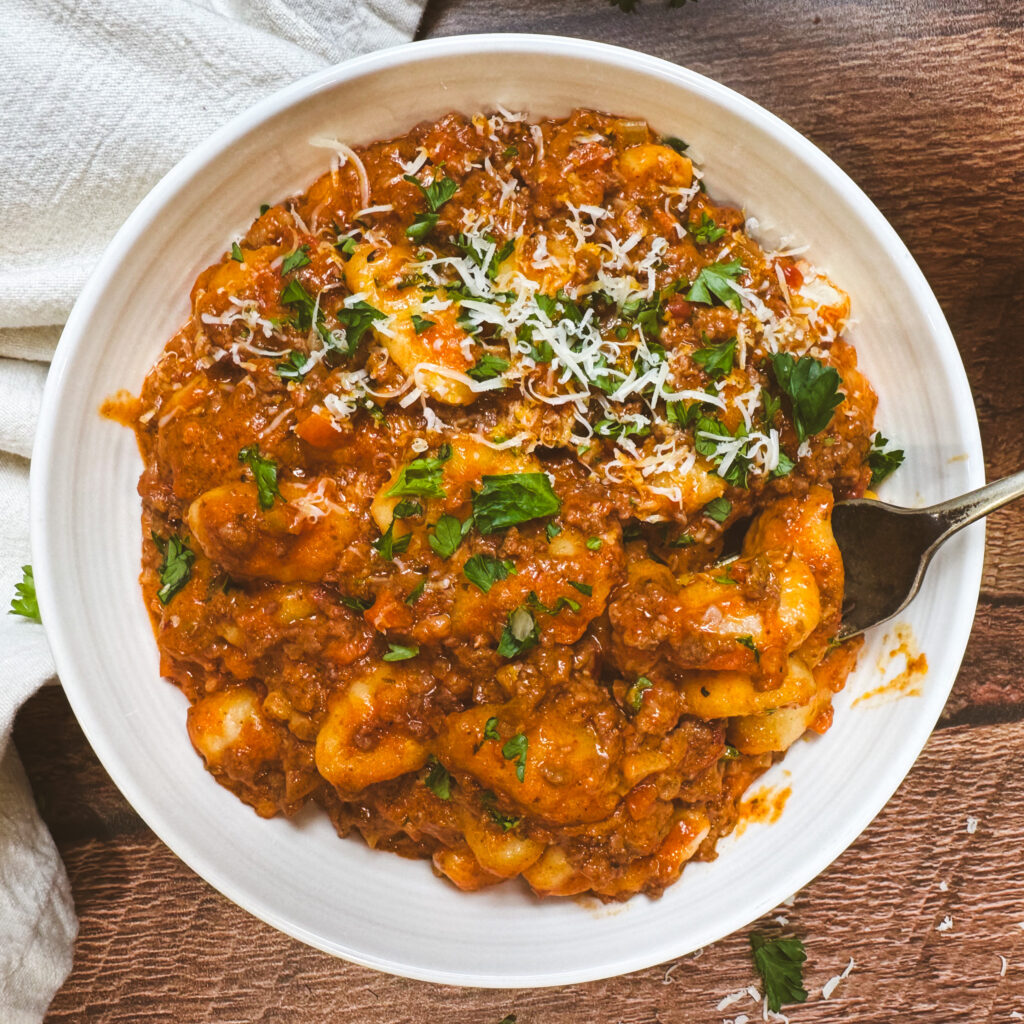 Finished one pot gnocchi bolognese topped with grated parmesan and fresh herbs in a bowl with a fork ready to eat.