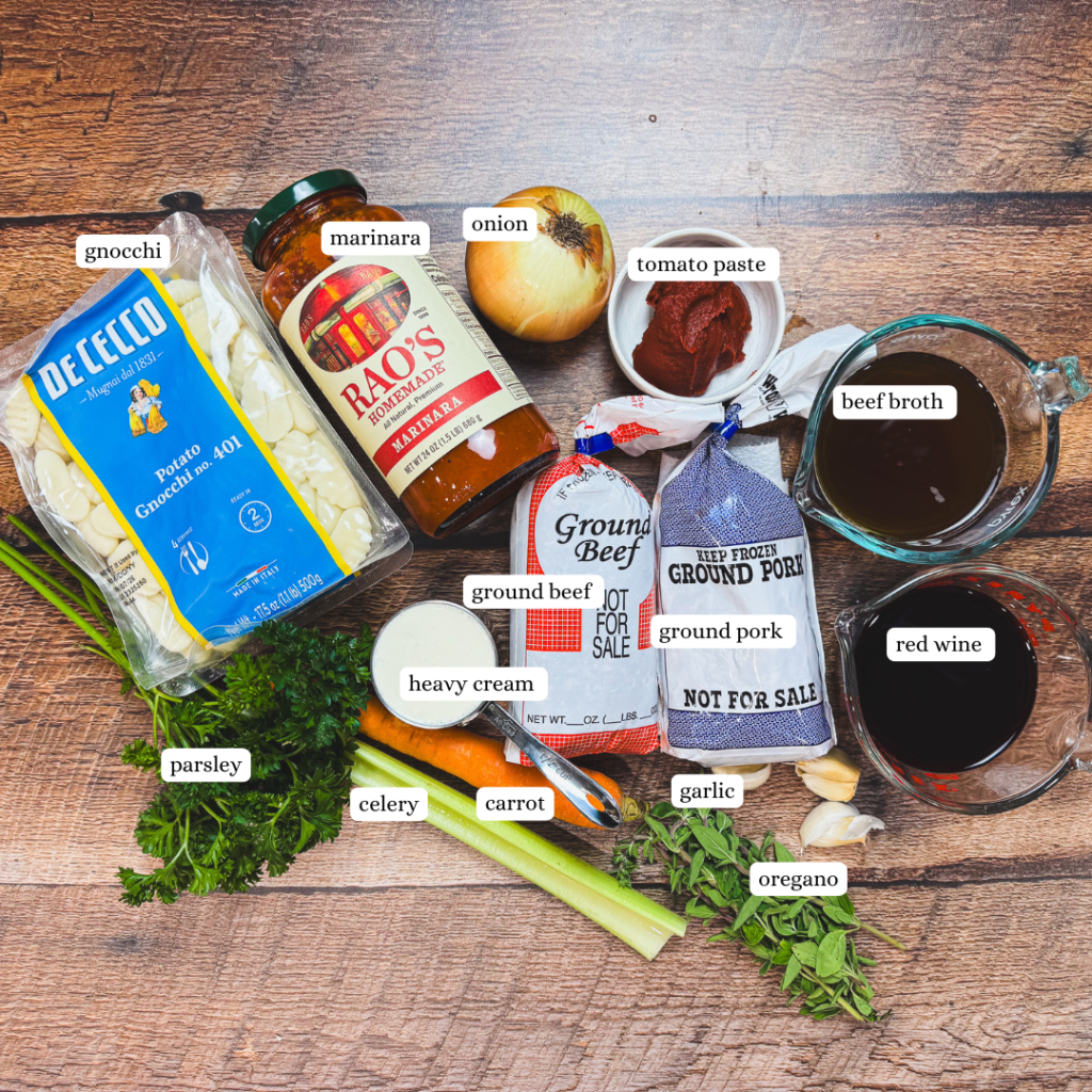 Ingredients for one pot gnocchi bolognese laid out on a wooden surface, including potato gnocchi, marinara sauce, ground beef, ground pork, onion, carrot, celery, garlic, tomato paste, broth, red wine, fresh herbs, and cream.