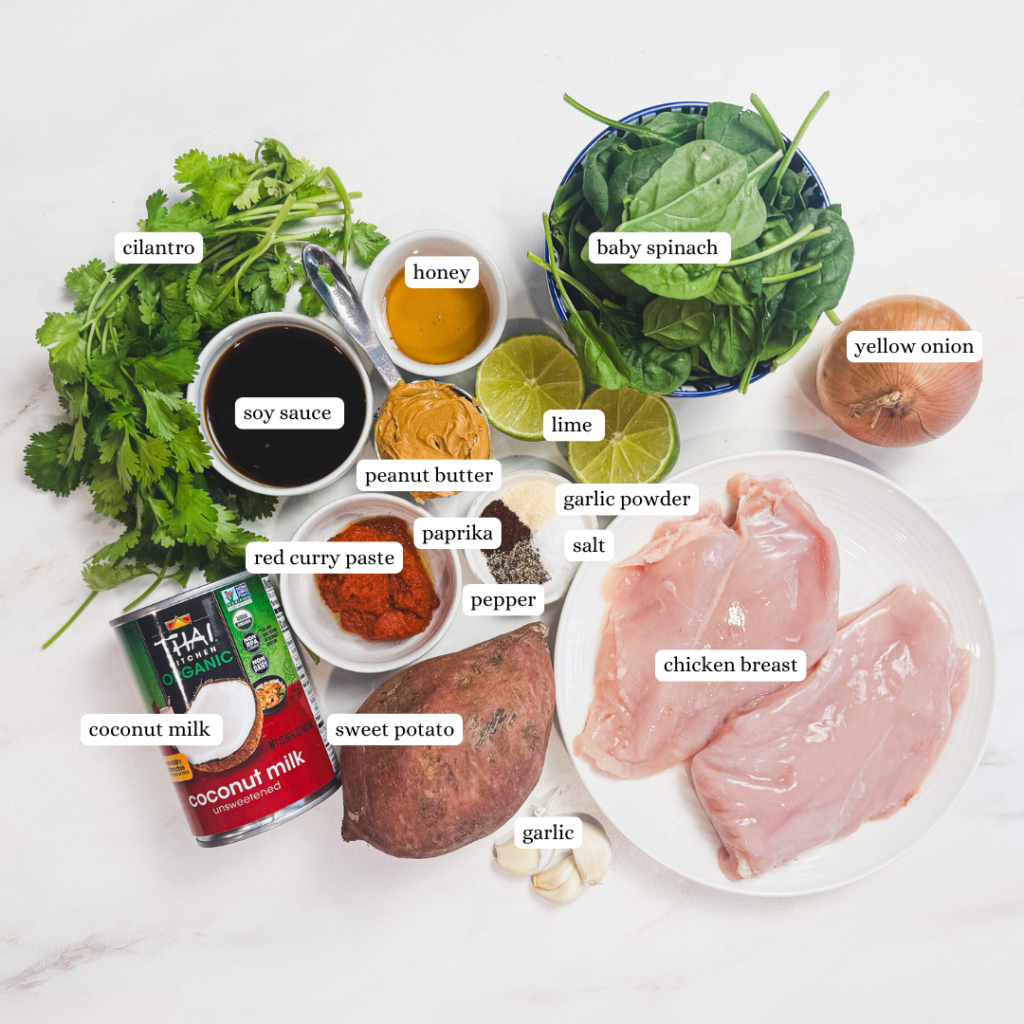 Flat lay of peanut chicken curry ingredients including raw chicken breasts, coconut milk, peanut butter, Thai red curry paste, sweet potato, spinach, lime, garlic, onion, cilantro, and spices on a white surface.