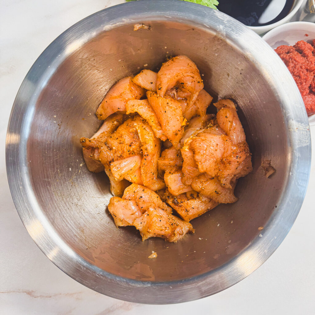 Raw chicken coated in spices and seasoning in a stainless steel mixing bowl.