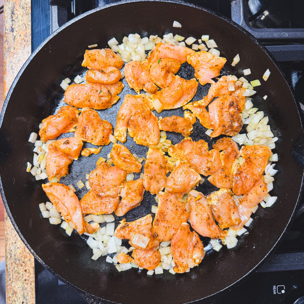 Seasoned chicken pieces browning in a cast iron skillet with diced onions.