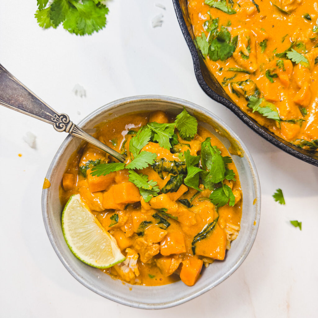 Bowl of peanut chicken curry with sweet potatoes, spinach, rice, fresh cilantro, and a lime wedge.