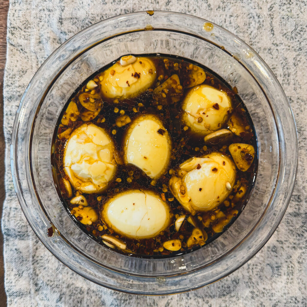 Jammy soft-boiled eggs marinating in soy sauce with garlic and red pepper flakes in a glass bowl.