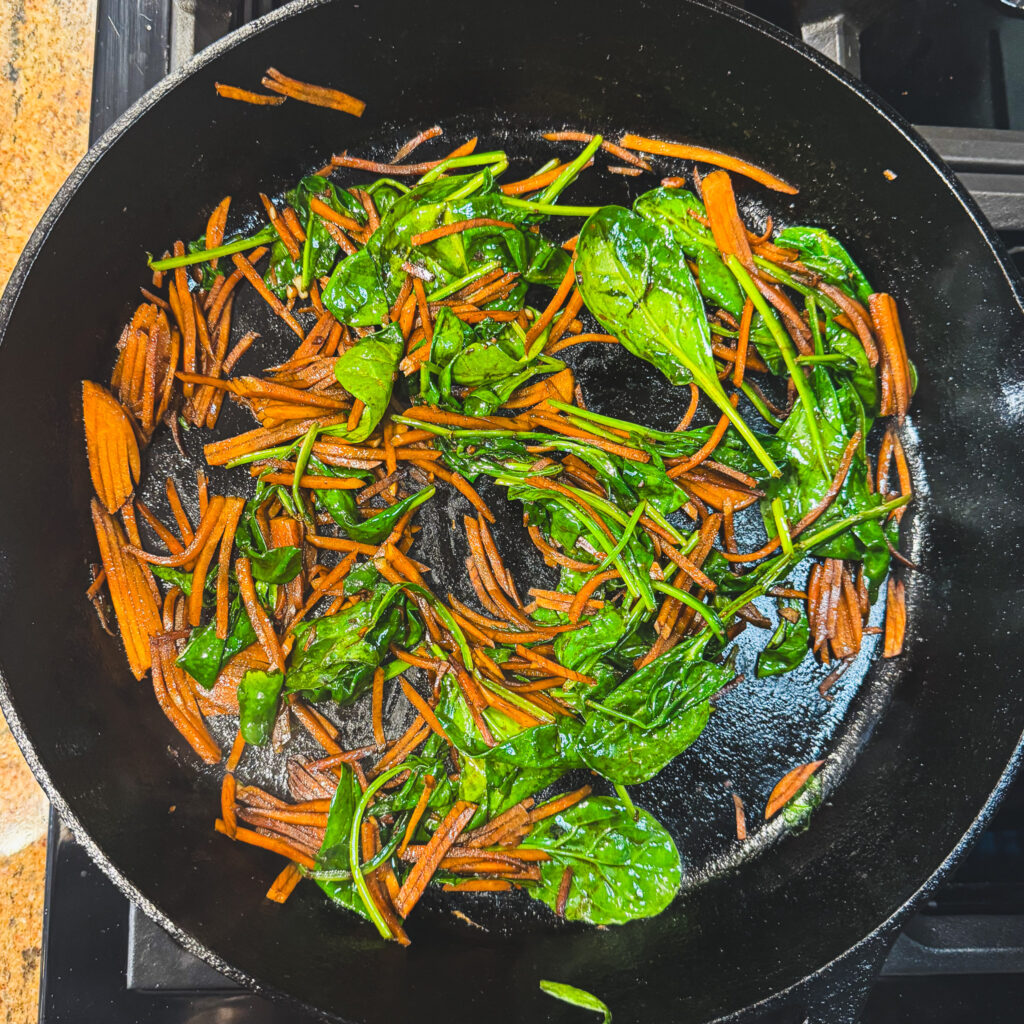 Sautéed carrots and baby spinach wilting together in a cast iron skillet.