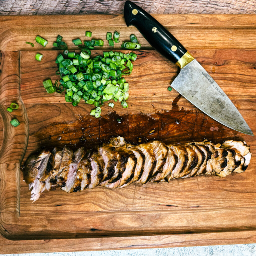 Sliced marinated pork tenderloin resting on a cutting board with chopped scallions.