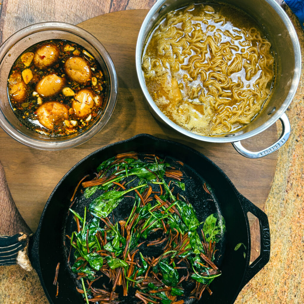 Prepared ramen components including soy eggs, cooked noodles in broth, and sautéed vegetables.