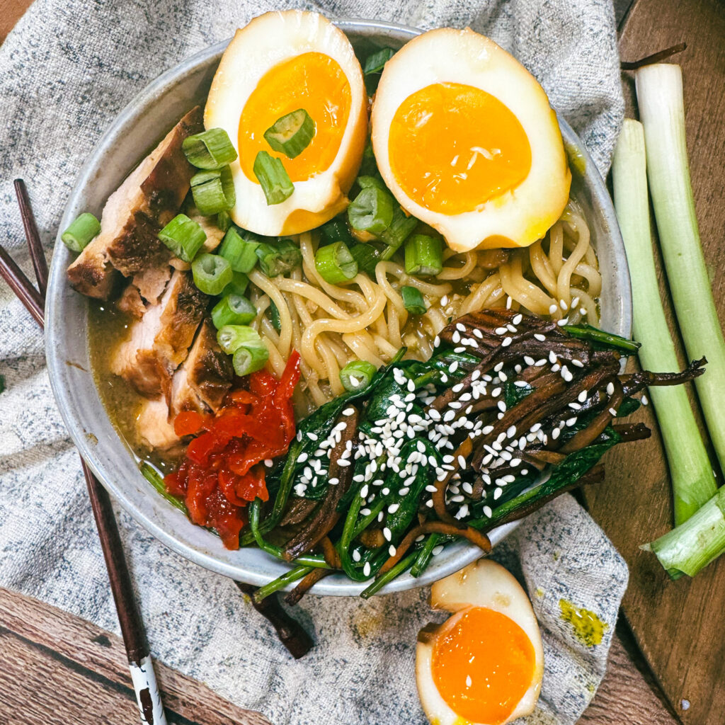 Overhead view of pork tenderloin ramen topped with jammy soy eggs, sliced pork, ramen noodles, sautéed spinach, mushrooms, scallions, sesame seeds, and chili paste.
