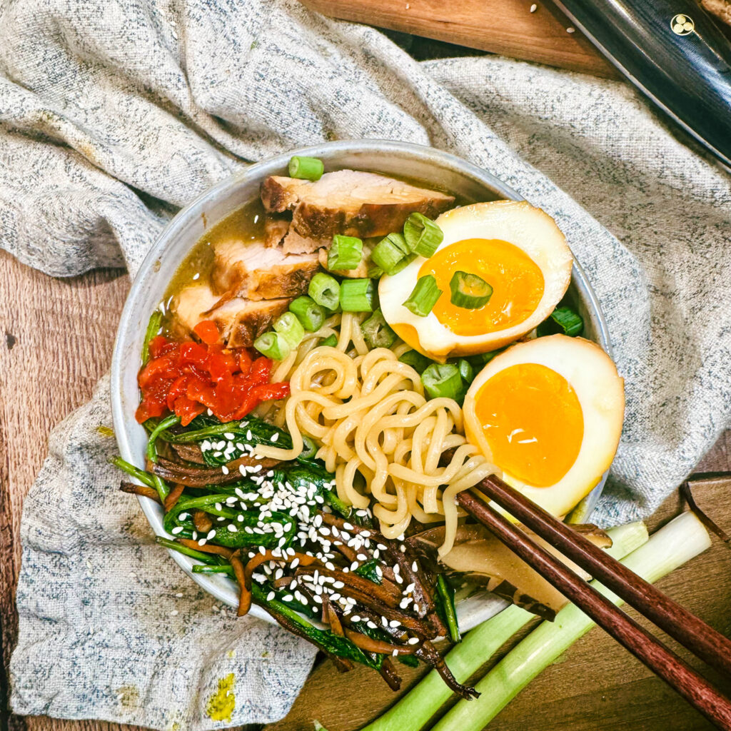 Pork ramen bowl with chopsticks lifting noodles, featuring soy eggs, marinated pork tenderloin, sautéed vegetables, and rich broth.
