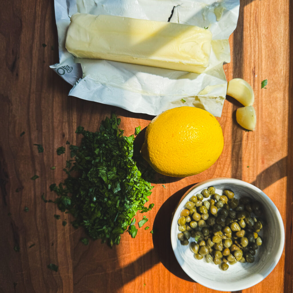 Overhead view of butter, fresh parsley, garlic, lemon, and capers on a wooden cutting board for making homemade lemon caper butter.