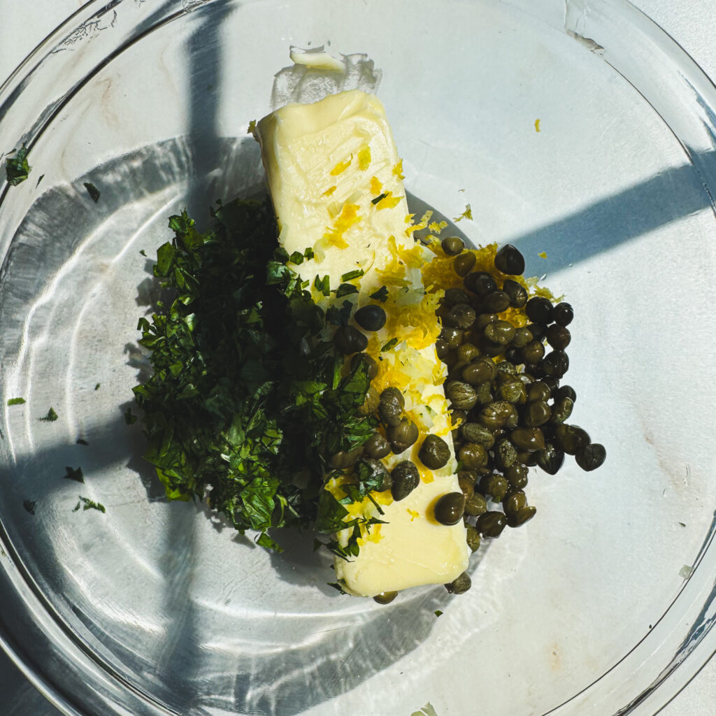 Butter topped with chopped parsley, lemon zest, and capers in a glass bowl before mixing into lemon herb compound butter.