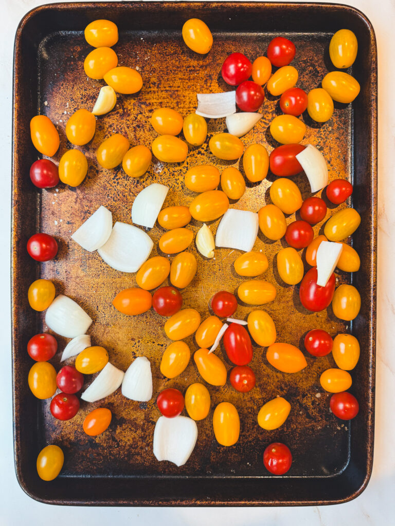 Overhead view of cherry tomatoes, onion, and garlic on a baking sheet before making homemade roasted tomato sauce.