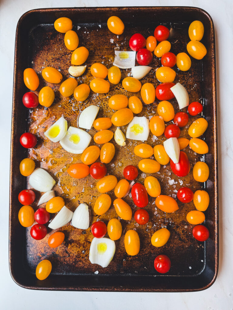 Cherry tomatoes, onion, garlic, olive oil, and flaky salt prepared on a sheet pan for roasted tomato sauce.