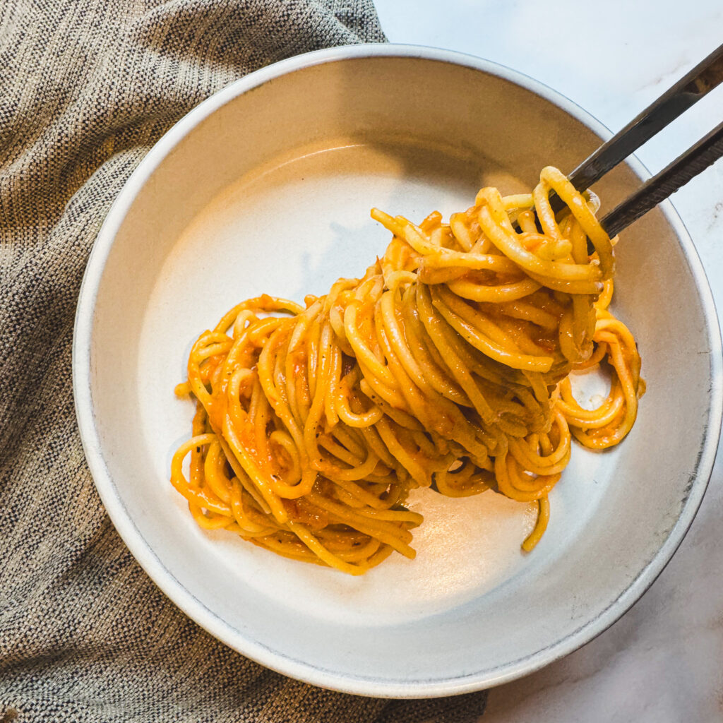 Creamy roasted tomato pasta twirled with tongs before serving in a shallow pasta bowl.