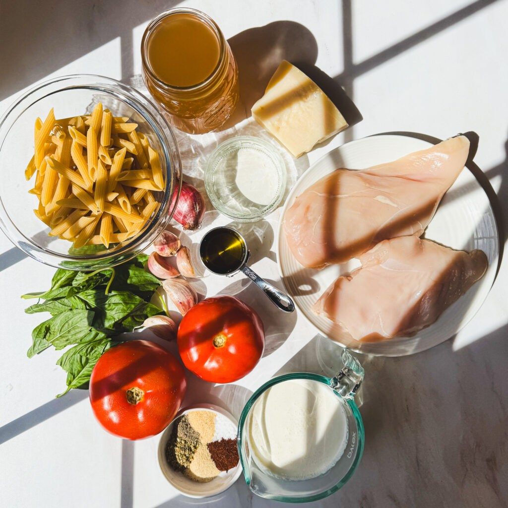Ingredients for one pot creamy bruschetta chicken pasta arranged on a white countertop with chicken, penne, tomatoes, basil, Parmesan, cream, broth, white wine, garlic, shallot, and seasonings.