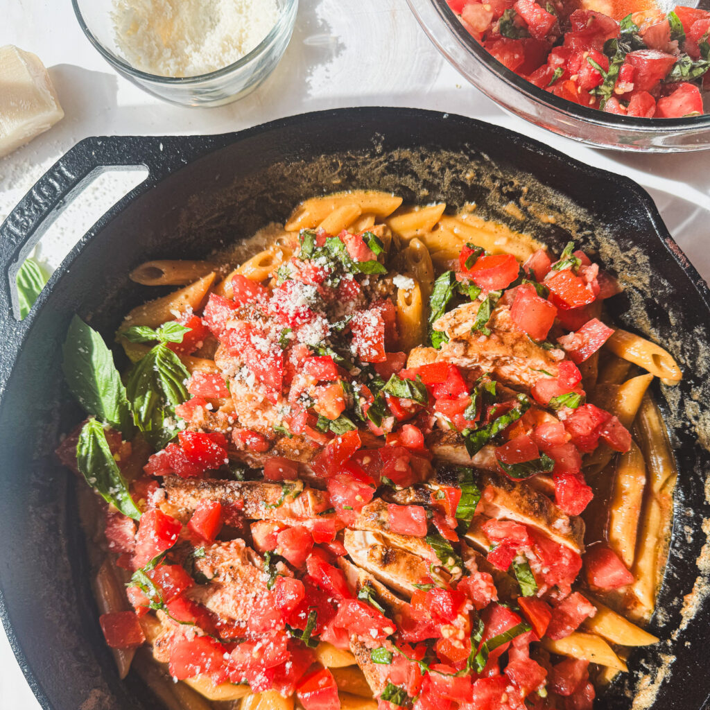 One pot creamy bruschetta chicken pasta garnished with fresh basil, homemade bruschetta, and Parmesan cheese in a cast iron skillet.