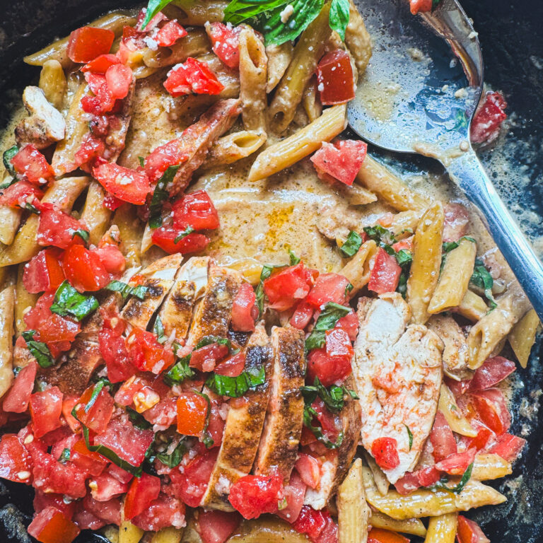 Creamy bruschetta chicken pasta in a cast iron skillet topped with fresh diced tomatoes, basil, Parmesan, and sliced seasoned chicken.