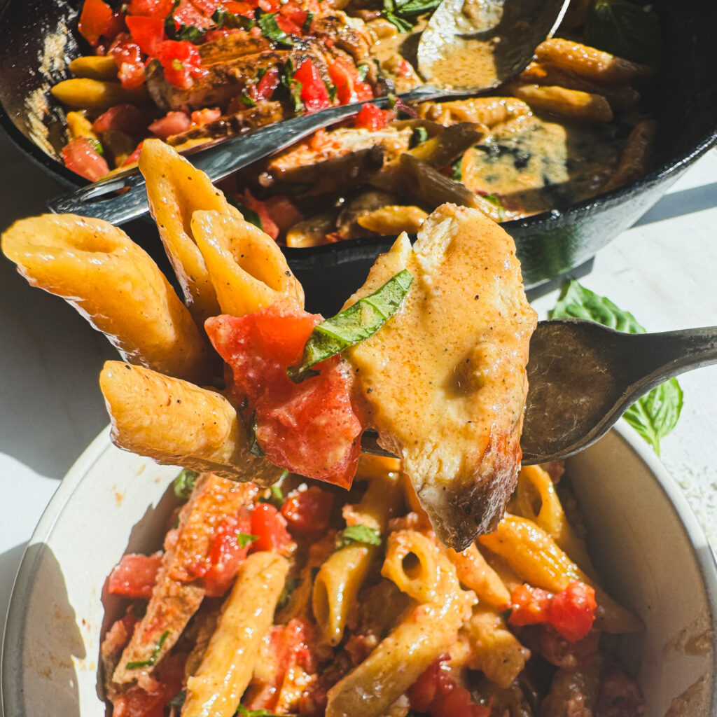 Close-up of a fork holding creamy penne pasta, seasoned chicken, fresh bruschetta tomatoes, and basil coated in a rich Parmesan cream sauce.