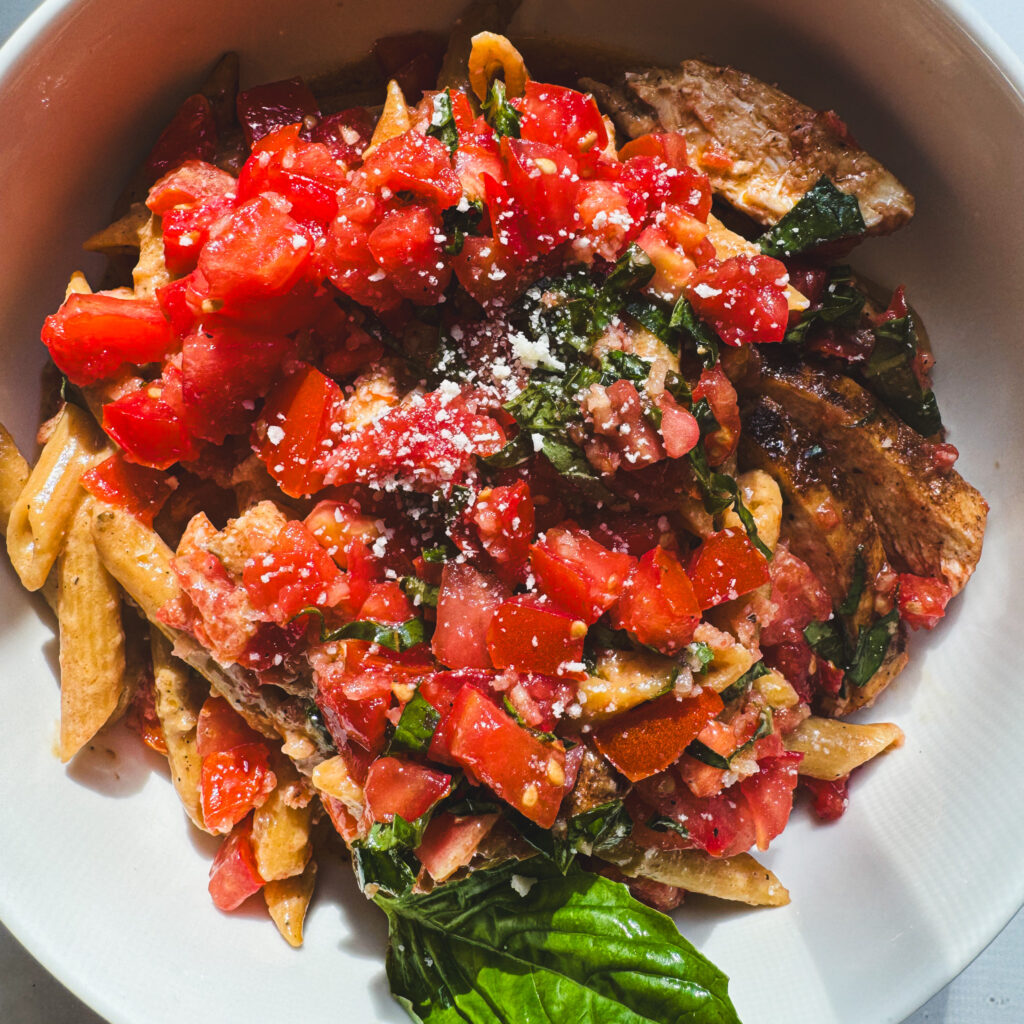 Bowl of creamy bruschetta chicken pasta topped with fresh tomato bruschetta, basil, grated Parmesan, and sliced chicken breast.