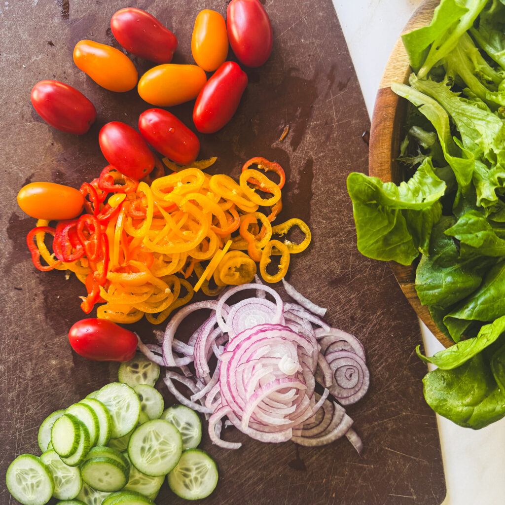Fresh salad ingredients including mixed greens, cherry tomatoes, mini sweet peppers, cucumber, and sliced red onion on a cutting board.