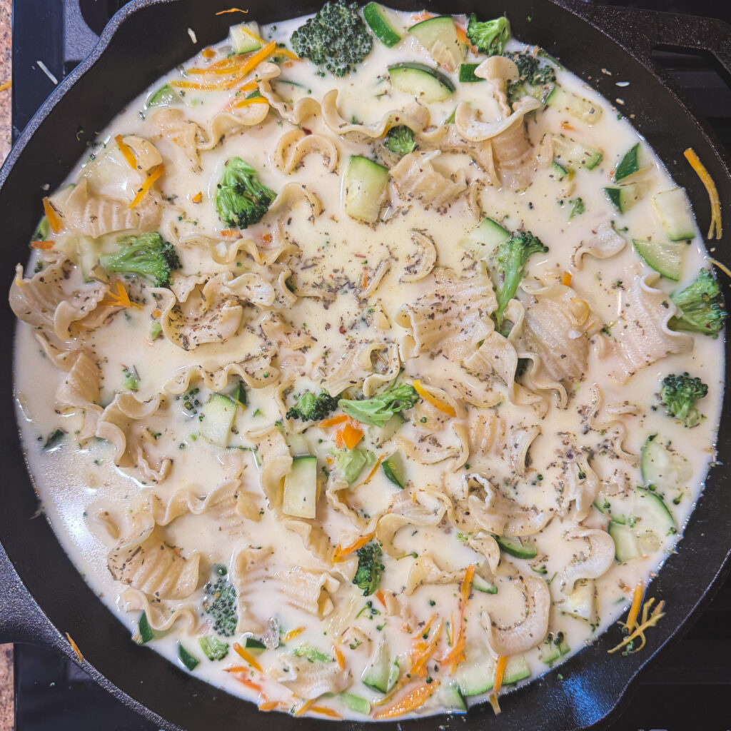 Wide lasagna-style noodles cooking with vegetables in a creamy white sauce in a cast iron skillet.