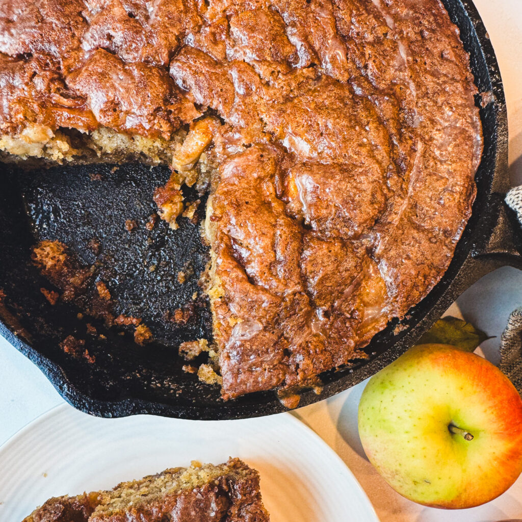 Slice removed: Cast iron apple fritter cake with a slice removed showing the moist brown butter cake and gooey apple filling.