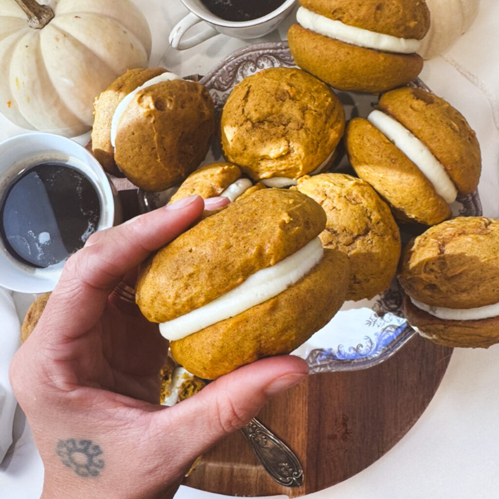 Hand holding a thick pumpkin whoopie pie filled with creamy cream cheese buttercream, surrounded by homemade whoopie pies and fall decorations.