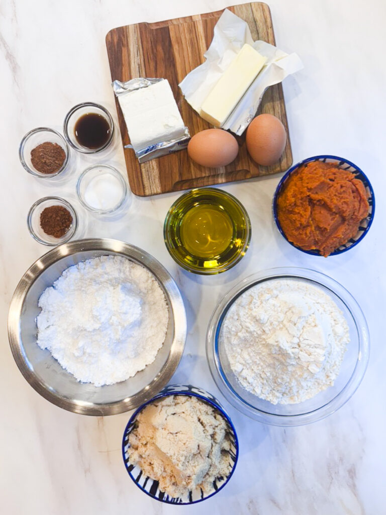 All ingredients for homemade pumpkin whoopie pies, including pumpkin puree, brown sugar, avocado oil, flour, eggs, butter, cream cheese, and spices.