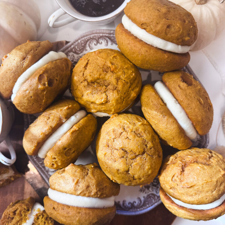 Homemade pumpkin whoopie pies filled with thick cream cheese buttercream, arranged on a vintage plate with white pumpkins and coffee.