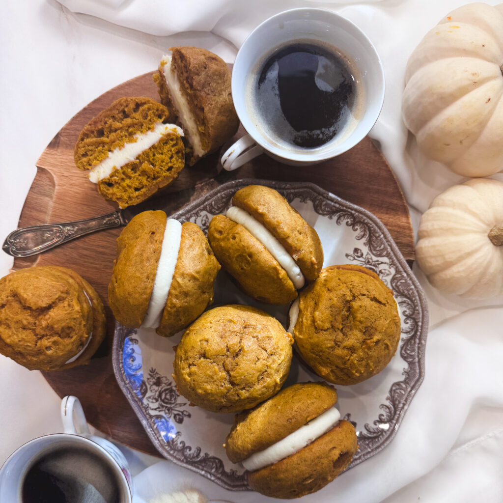 Homemade pumpkin whoopie pies with thick cream cheese filling, served on a vintage plate with decorative white pumpkins and coffee.