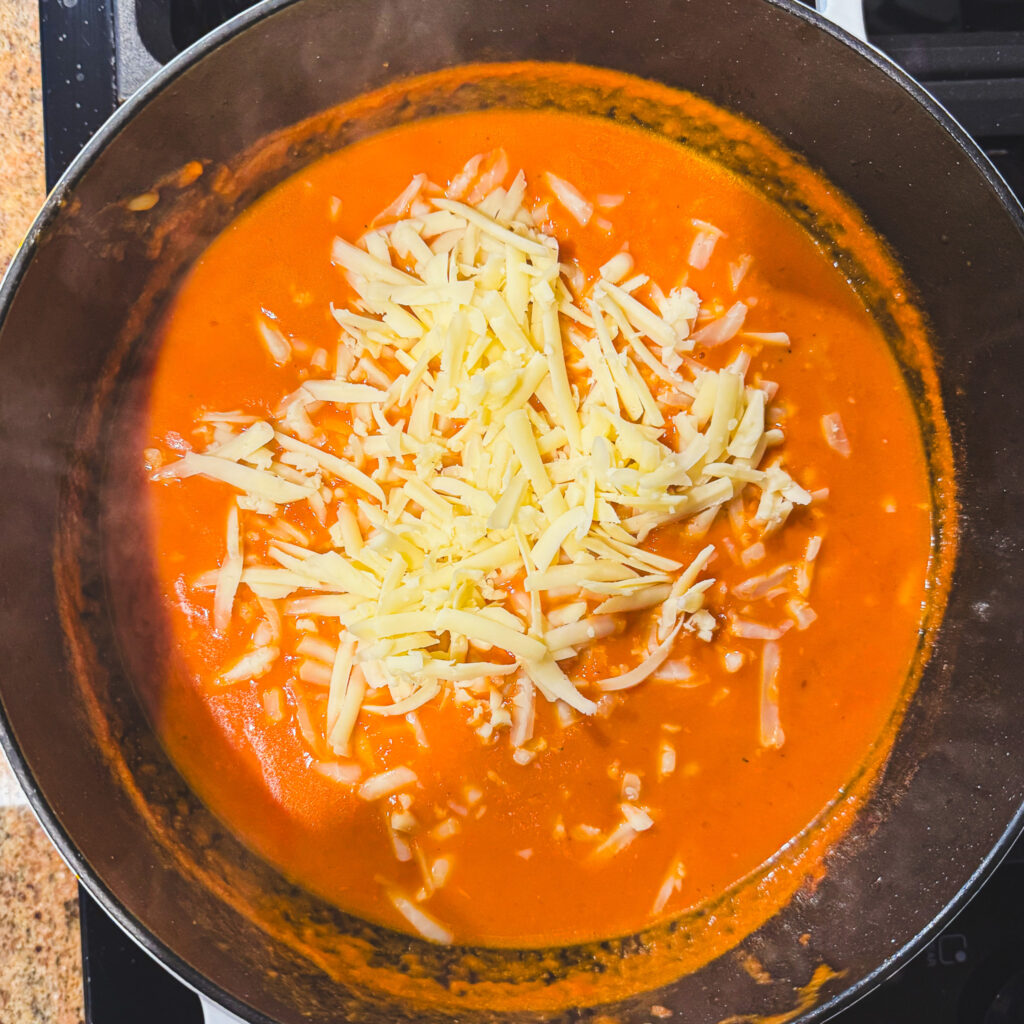 Adding cheddar: Freshly shredded cheddar cheese added to creamy tomato soup in a Dutch oven.